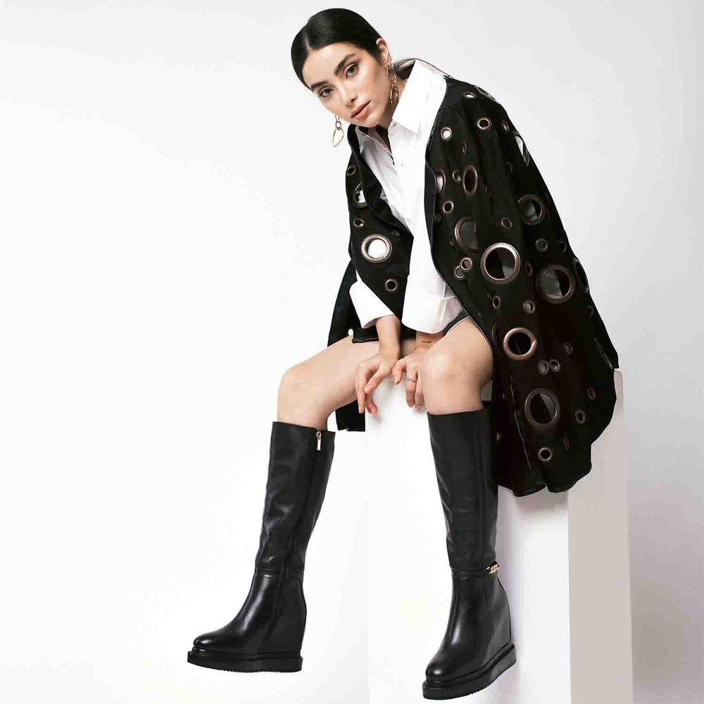 Model wearing ladies black leather wedge knee-high boots with chain detail