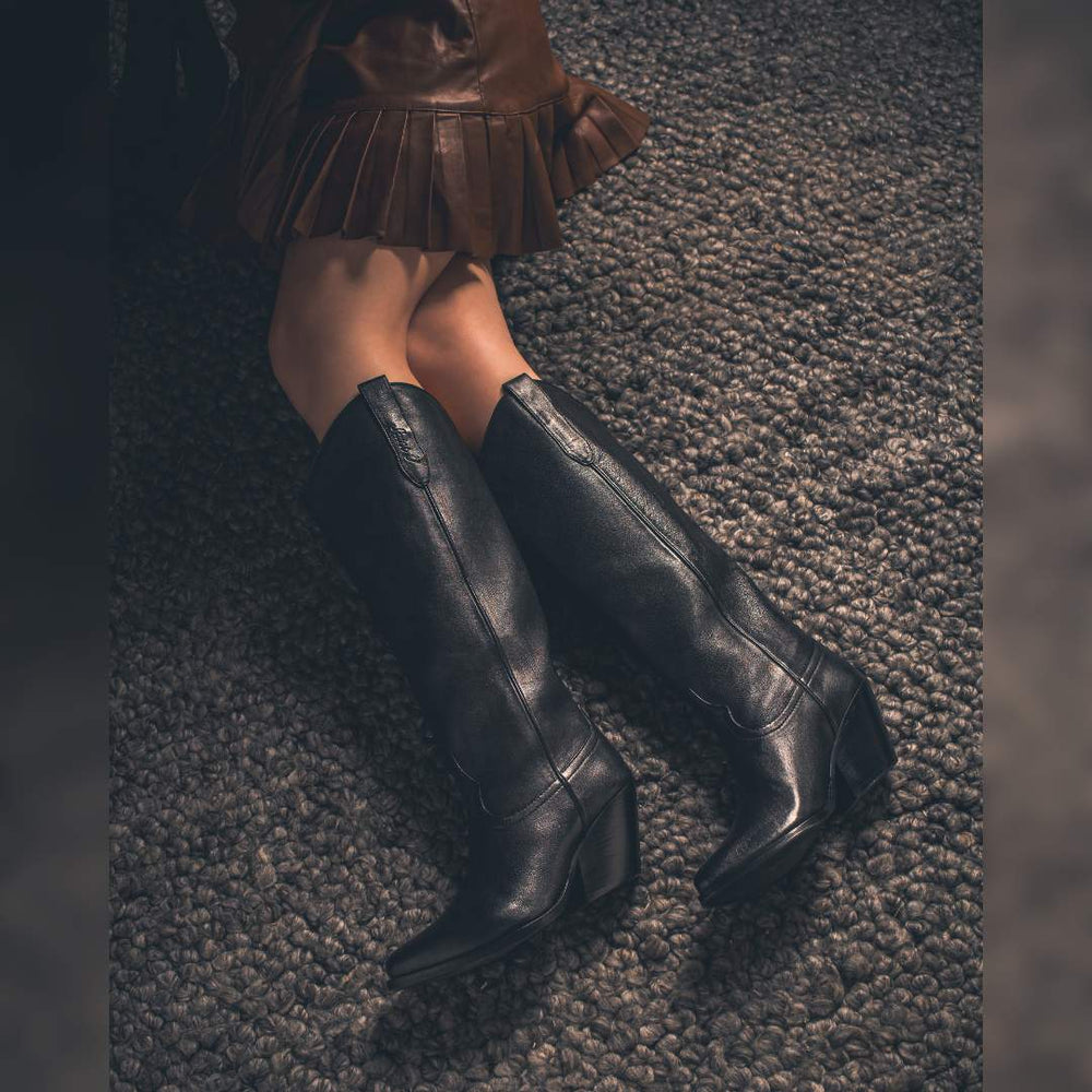 Women's black leather knee high boots by Saint Loanna, styled seated lifestyle shot with rust-toned skirt